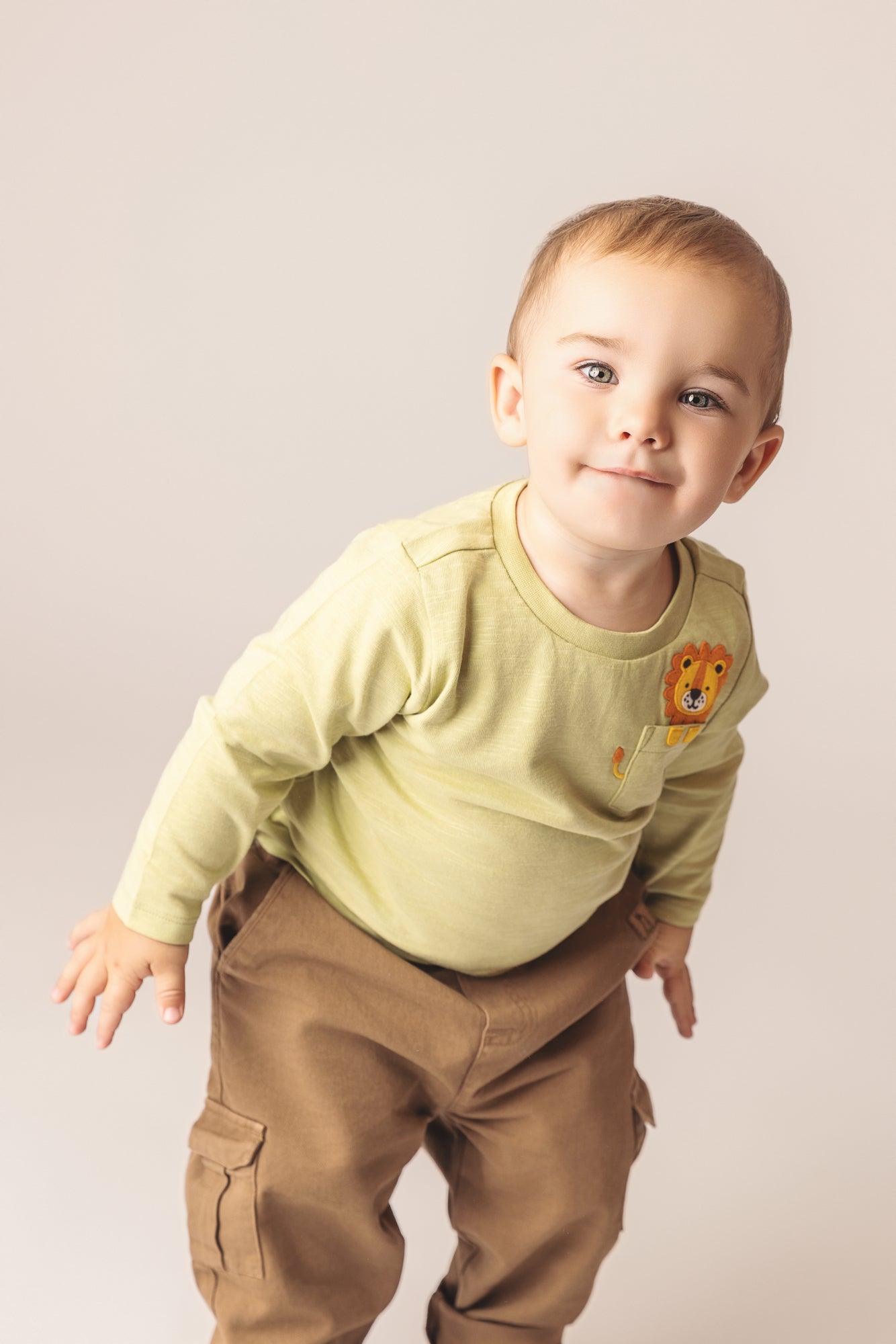Baby in a light olive green long-sleeved shirt with a lion embroidered on the pocket wearing brown cargo pants.