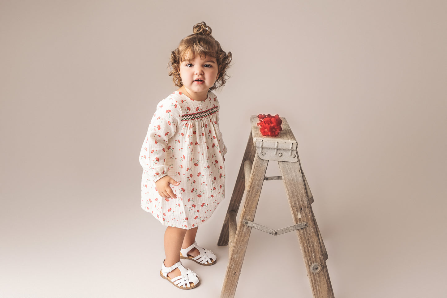Baby girl in a floral dress wearing white sandals and standing next to a wooden stepladder with red flowers.