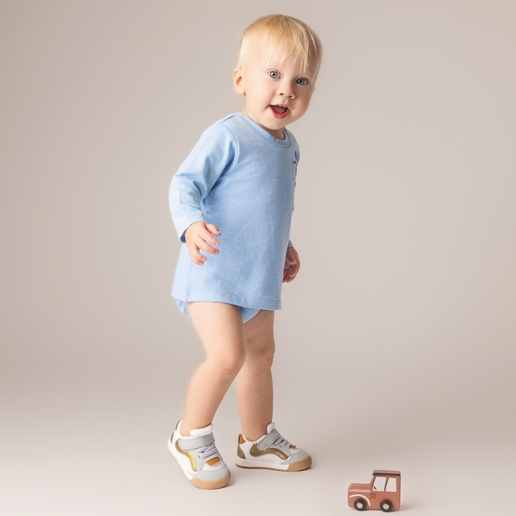 Baby in light blue long-sleeve outfit and sneakers, smiling, standing next to a small toy car.