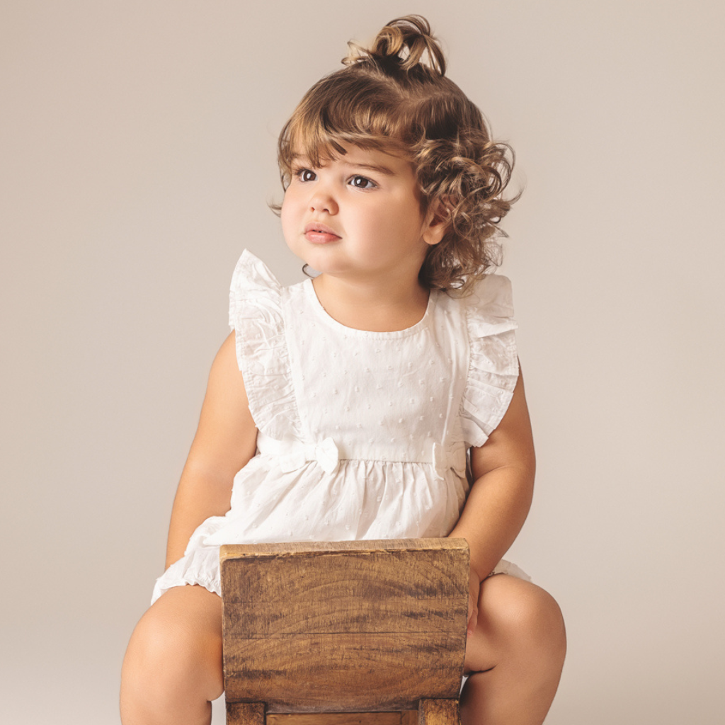 Baby girl in a white eyelet dress with ruffled sleeves sits on a wooden stool.