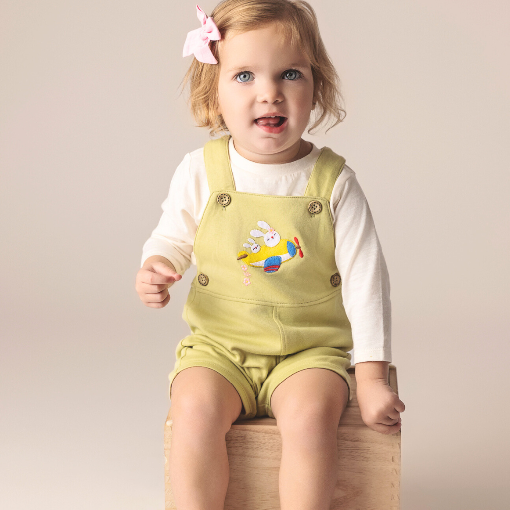 Baby girl wearing light yellow overalls with embroidered airplane and bunnies.