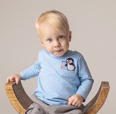 Baby boy in light blue shirt with penguin patch wearing gray cargo pants and white shoes seated on wooden chair.