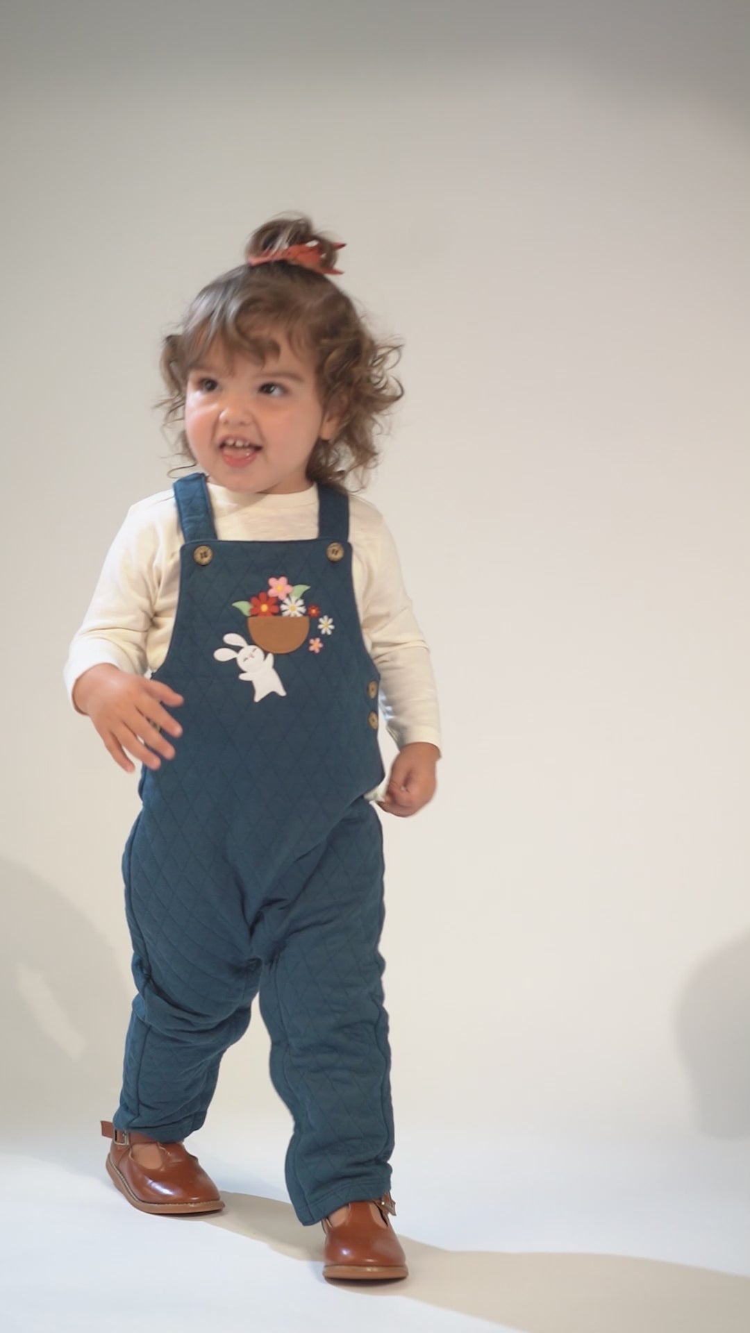 Toddler in teal quilted overalls with a bunny and flower embroidery, wearing brown leather shoes.