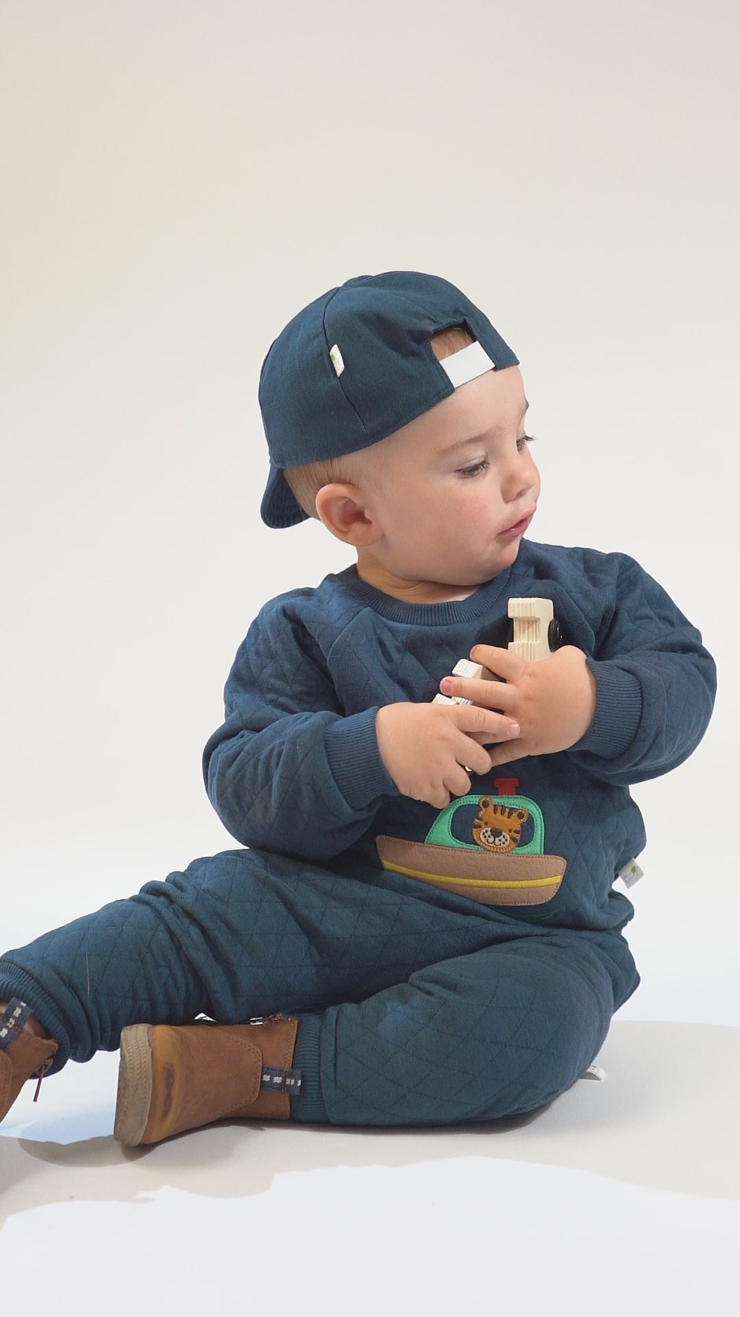 Baby in a teal sweatshirt and pants with a tiger-ship applique, navy baseball cap, and brown boots.